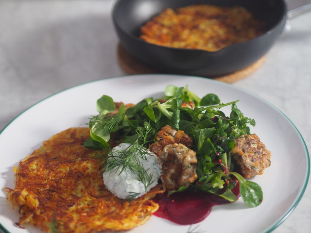 Ein Teller mit Röstkartoffeln und Seesaibling und Salat dahinter eine Pfanne mit noch mehr Rösti