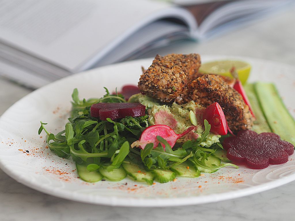 Lachs im Chia-Knuspermantel mit Gurkenstreifen und Avocado-Püree auf einem Teller im Hintergrund das Kochbuch, aufgeschlagen.
