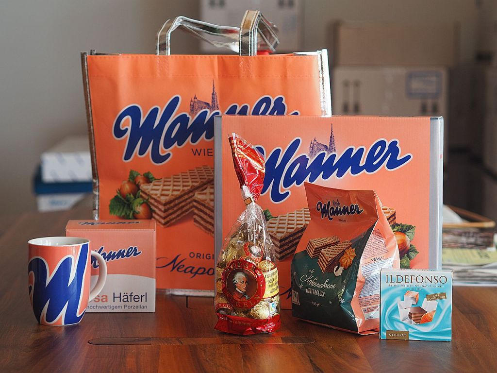 Ein Tisch mit verschiedenen Produkten der Marke Manner, einschließlich einer Papiertüte, Waffeln, Schokolade, einem Kaffeebecher und einer Verpackung mit Nougat.