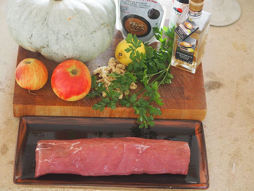 Eine Auswahl von frischen Zutaten für eine Mahlzeit, einschließlich eines grauen Kürbisses, roter Äpfel, Walnüsse, frischer Petersilie, einer Zitrone, hochwertigem Öl und einem Stück rotem Fleisch auf einem dunklen Teller.