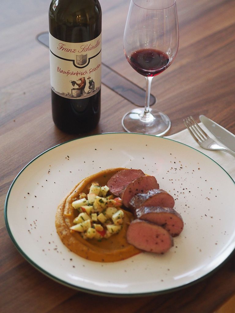 Ein Teller mit Scheiben von zartem Fleisch auf einer Kürbispüree-Basis, garniert mit einer frischen Apfelbeilage, daneben ein Glas Rotwein und eine Flasche Blaufränkisch.