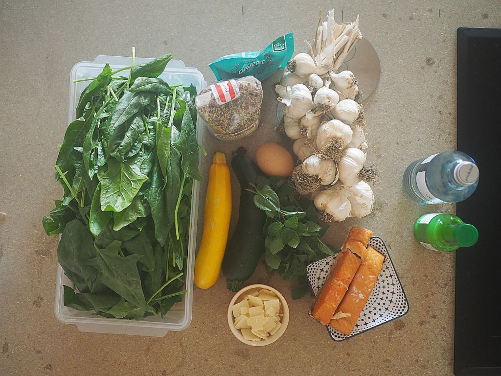 Eine Anordnung frischer Zutaten auf einer Arbeitsfläche, darunter Spinat, Zucchini, gelbe Zucchini, Knoblauch, ein Ei, frische Kräuter, zwei Stangen Brot, geriebener Käse, Wasserflasche und ein grünes Getränk.