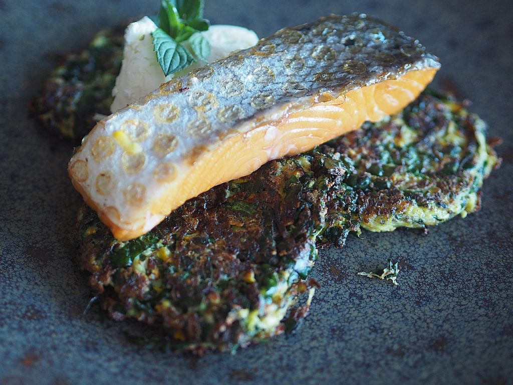 Ein Gericht mit gebratenem Lachsfilet auf zwei Gemüse-Pfannkuchen, garniert mit frischer Minze und einem Klecks Creme.