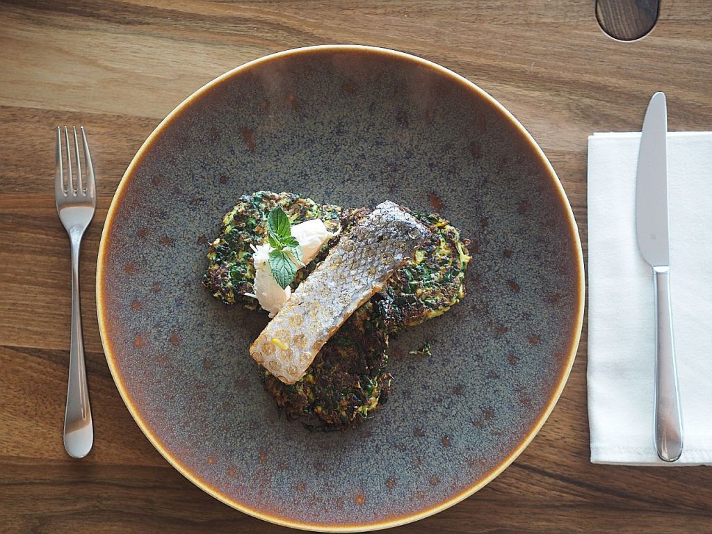 Ein Teller mit gebratenem Fischfilet, serviert auf einem Bett aus Gemüse-Patties, garniert mit einem Klecks Sahne und frischer Minze.