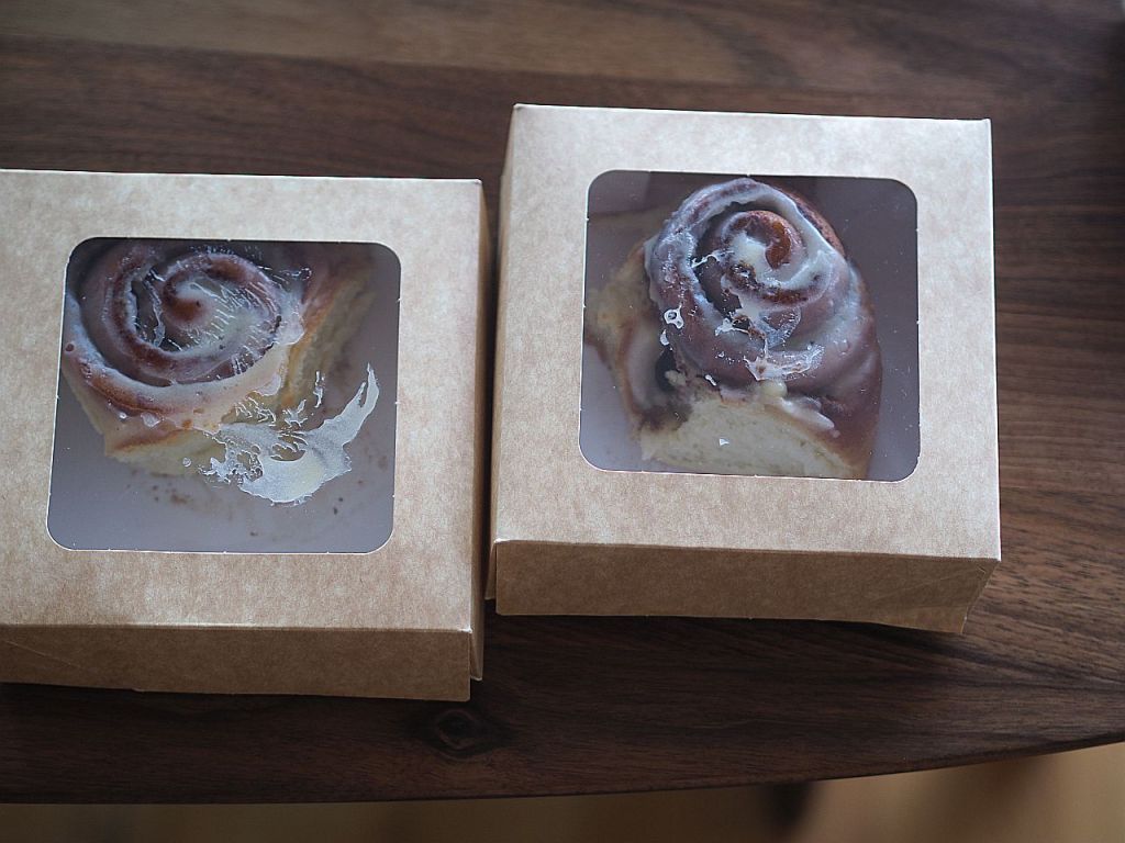 Zwei verpackte Zimtschnecken in durchsichtigen Boxen auf einem Holzuntergrund.
