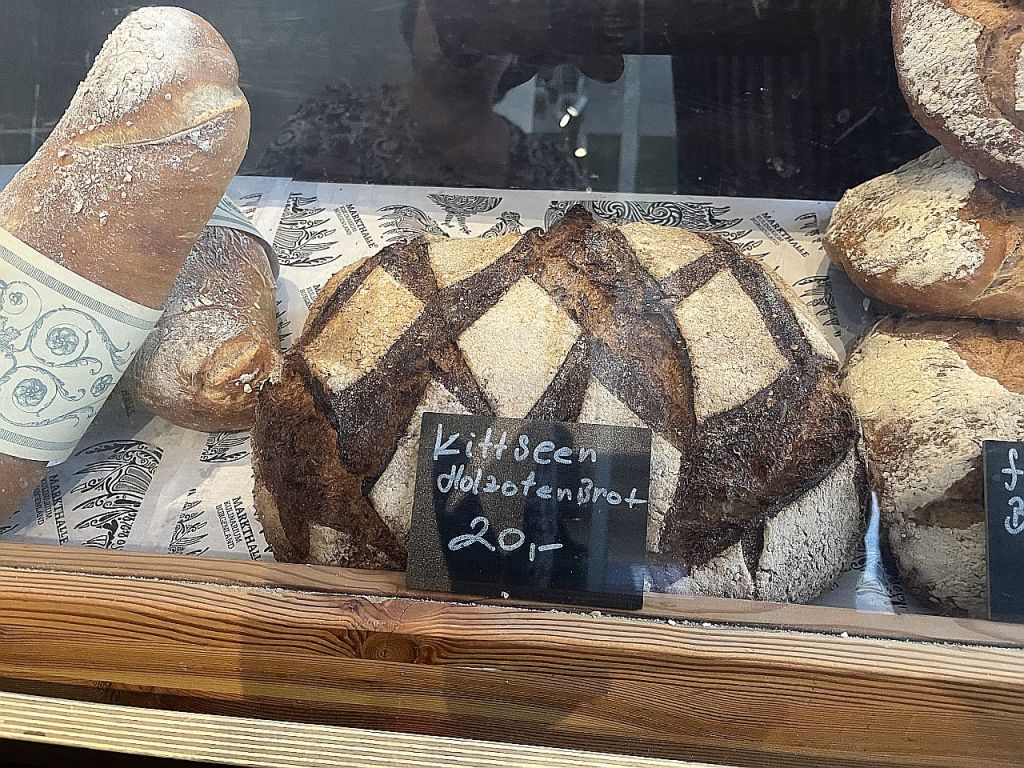 Ein Holzofenbrot mit einer rautenförmigen Kruste, beschriftet mit "Kittseen Holzenbrot" und einem Preis von 20 Euro, zusammen mit anderen Broten in einer Bäckereiauslage.