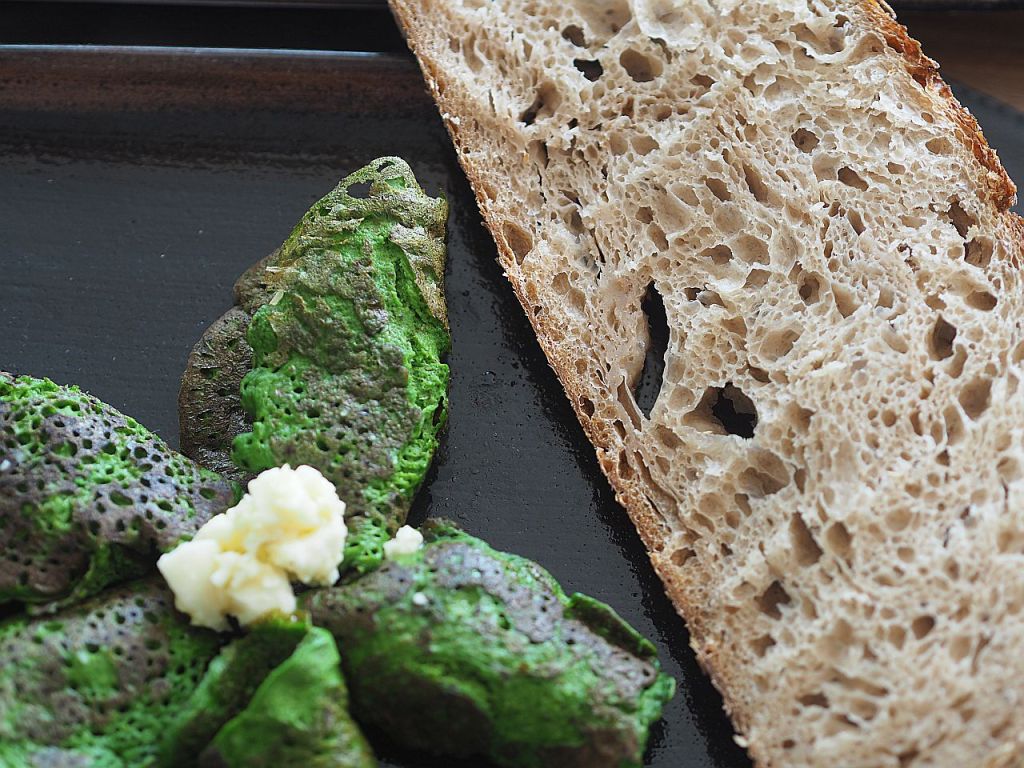 Ein Teller mit grünlichen, unregelmäßig geformten essbaren Stückchen und einem Stück braunem Brot, serviert auf schwarzem Geschirr.