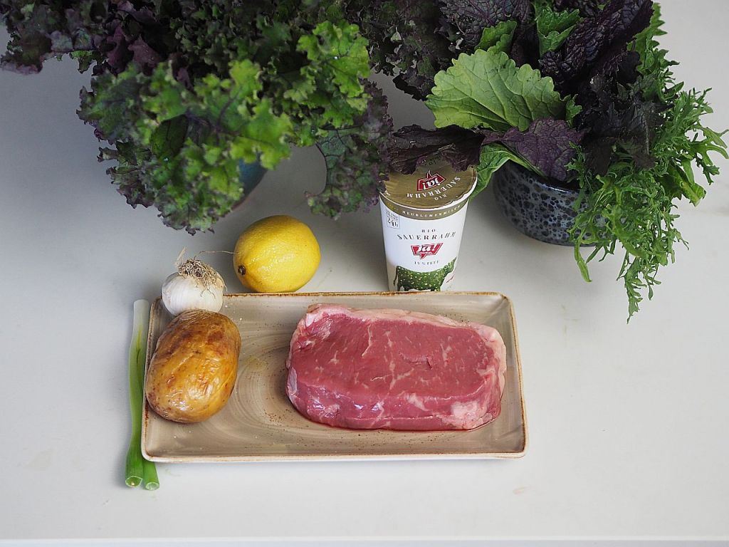 Eine Platte mit rohem Steak, einer Kartoffel, einer Zitrone, Knoblauch und Frühlingszwiebeln, umgeben von verschiedenen grünen Blättern und einem Becher Sauerkraut.