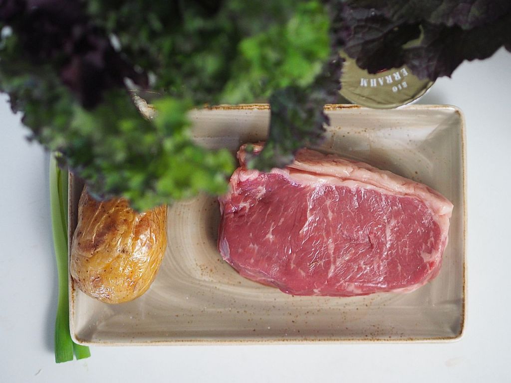 Ein Stück rotes Fleisch liegt auf einem Teller, daneben eine gebackene Kartoffel und einige grüne Blätter von Gemüse.