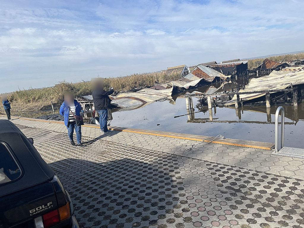 Zwei Personen stehen auf einem Steg neben einem Wasserbereich mit überfluteten, beschädigten Strukturen im Hintergrund. Ein Auto ist im Vordergrund sichtbar.