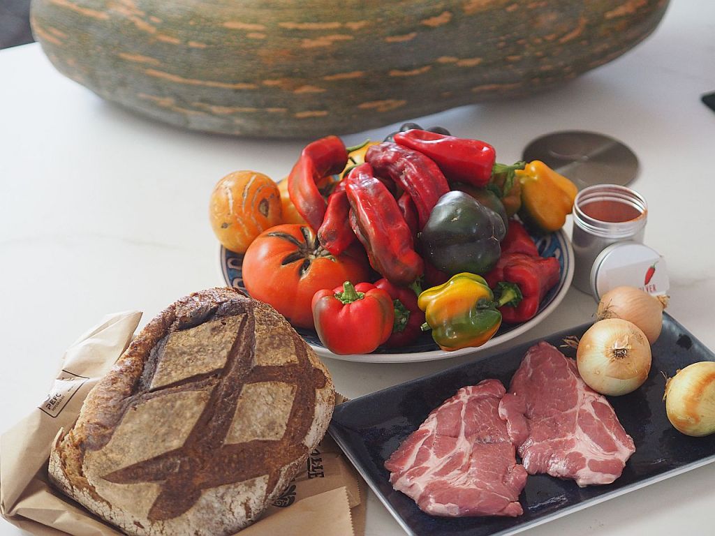 Ein Tisch voller frischer Zutaten, darunter eine Auswahl an buntem Gemüse wie Paprika und Tomaten, geschnittenes Fleisch auf einem dunklen Teller, eine Kruste Brot und Zwiebeln, mit einem großen Kürbis im Hintergrund.