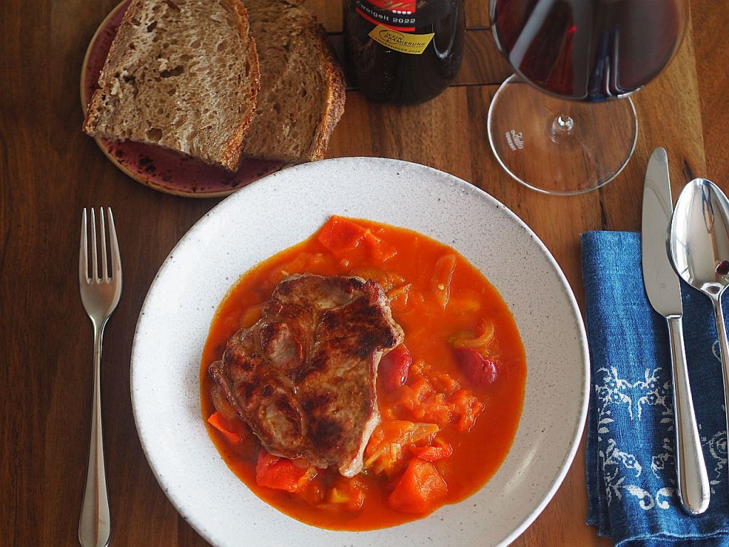 Ein Teller mit Fleisch in einer bunten Sauce, umgeben von Gemüse, serviert mit einer Scheibe dunklem Brot, einem Glas Rotwein und Besteck auf einem Holztisch.