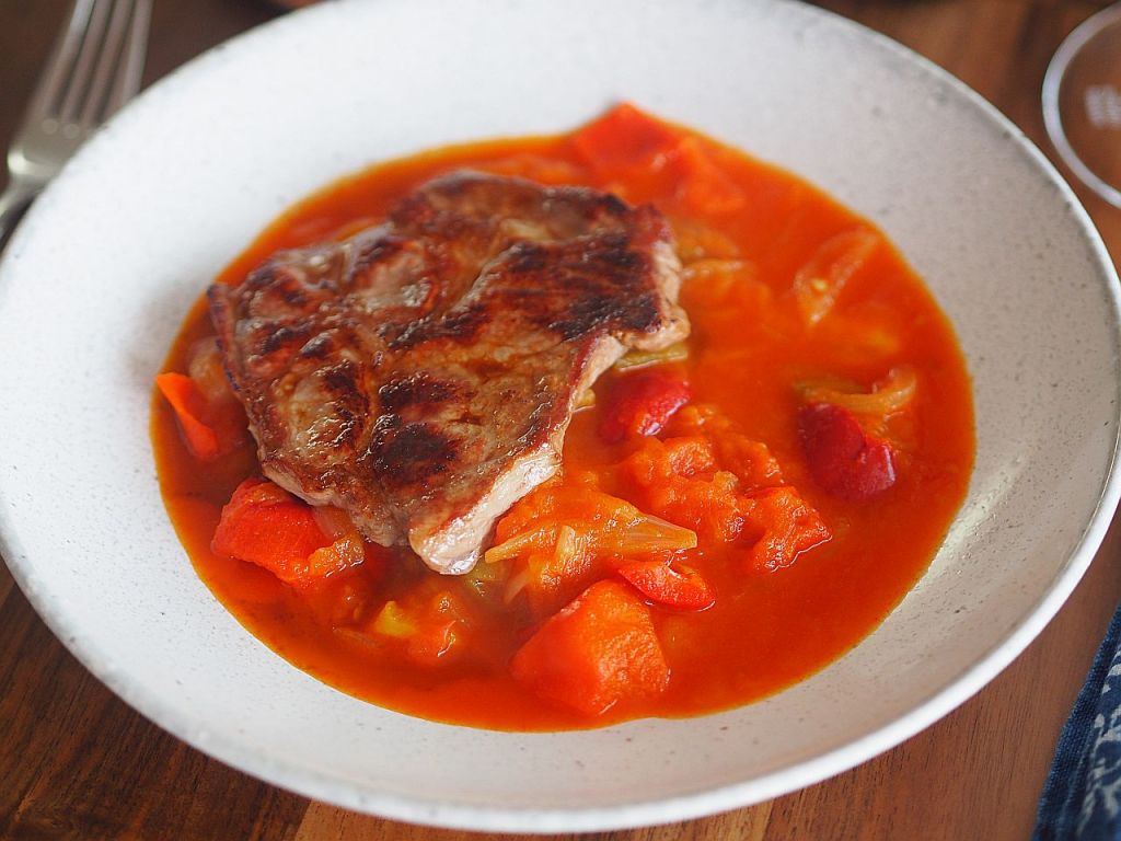 Ein Teller mit einem Stück gegrilltem Fleisch, umgeben von einer bunten Gemüsesauce mit Karotten, Paprika und Tomaten.