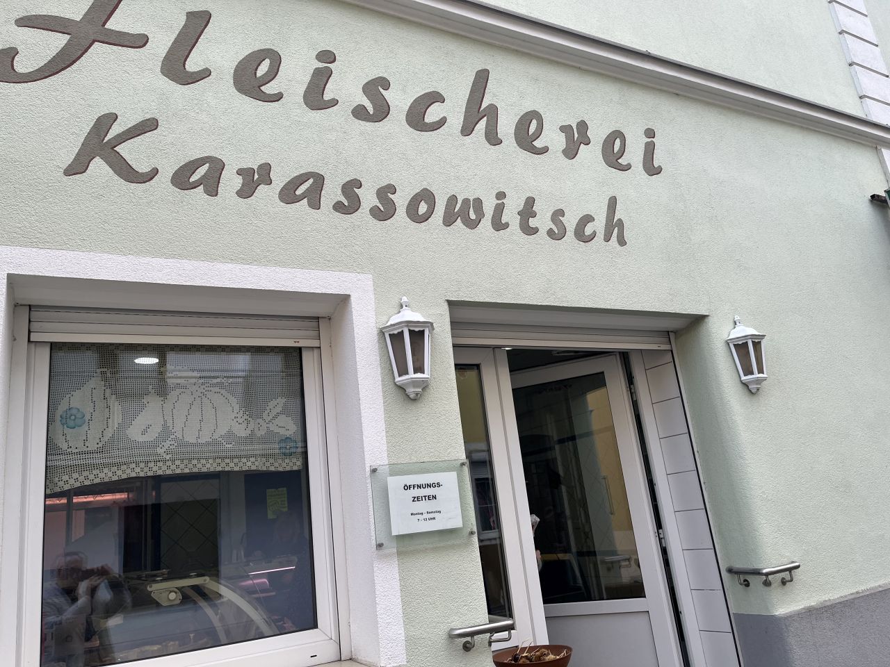 Außenansicht einer Fleischerei namens 'Karassowitsch' mit einem grünen Fassadenanstrich und einem Fenster mit Dekorvorhängen.