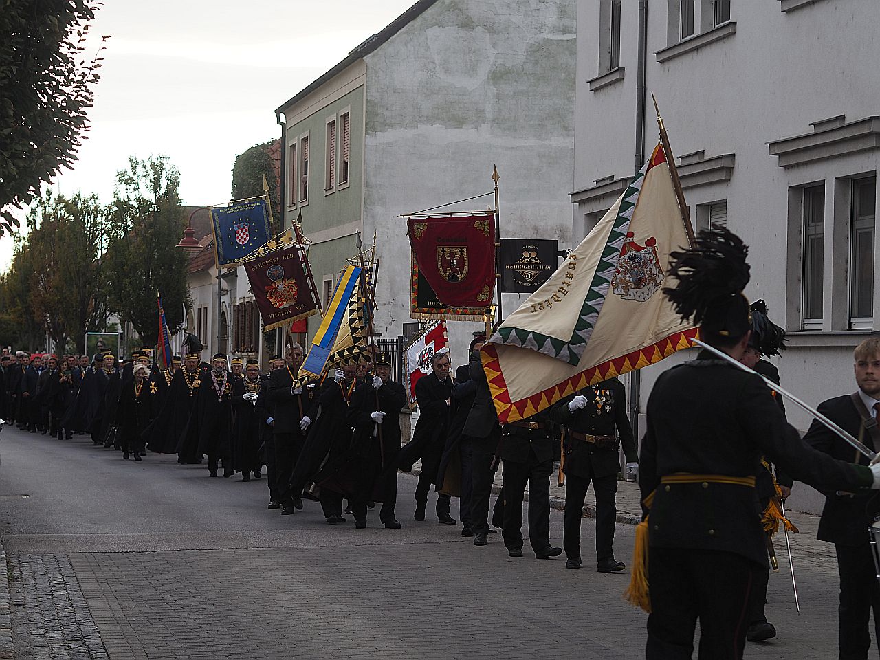 Eine festliche Parade mit Teilnehmern in schwarzen Umhängen, die Fahnen und Banner tragen, zieht durch eine Straße, flankiert von Gebäuden und Bäumen.
