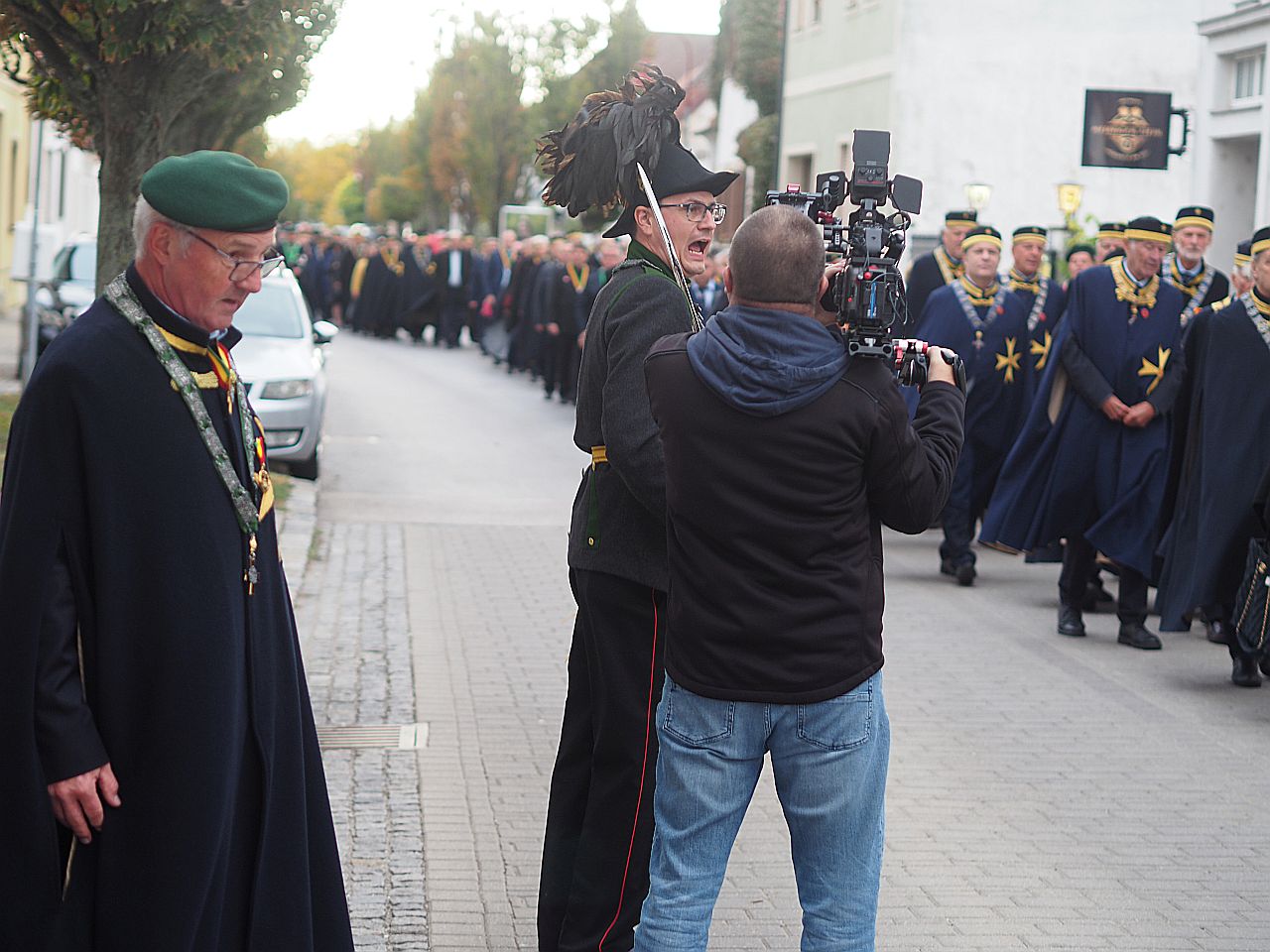 Eine Gruppe von festlich gekleideten Menschen marschiert durch eine Straße, während ein Kameramann einen Redner filmt. Im Vordergrund sehen wir zwei Männer in traditioneller Kleidung mit Medaillen.