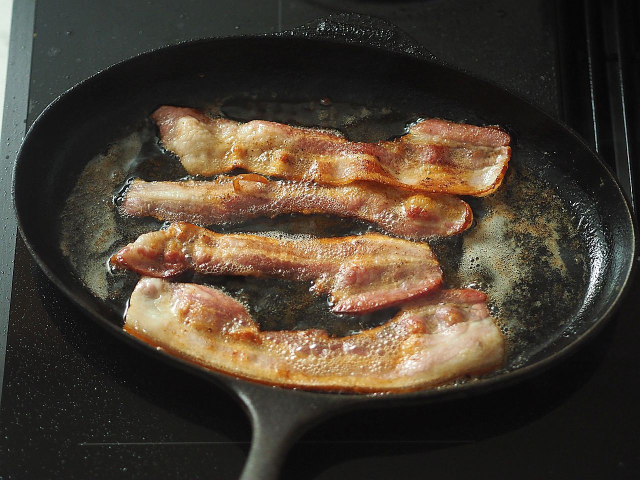 Scheiben von Speck braten in einer gusseisernen Pfanne auf einem Herd.