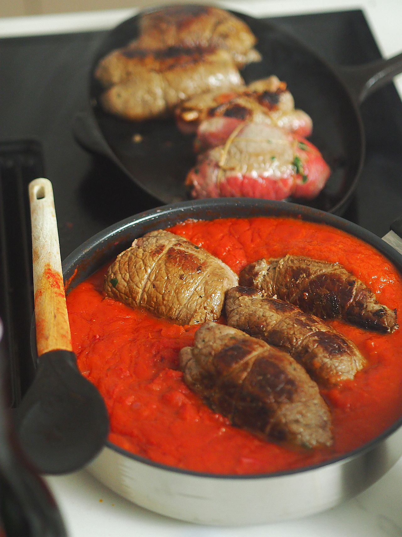 Ein Topf mit gefülltem Fleisch, das in einer roten Tomatensauce kocht, neben einer Pfanne mit weiteren gegrillten Fleischstücken.