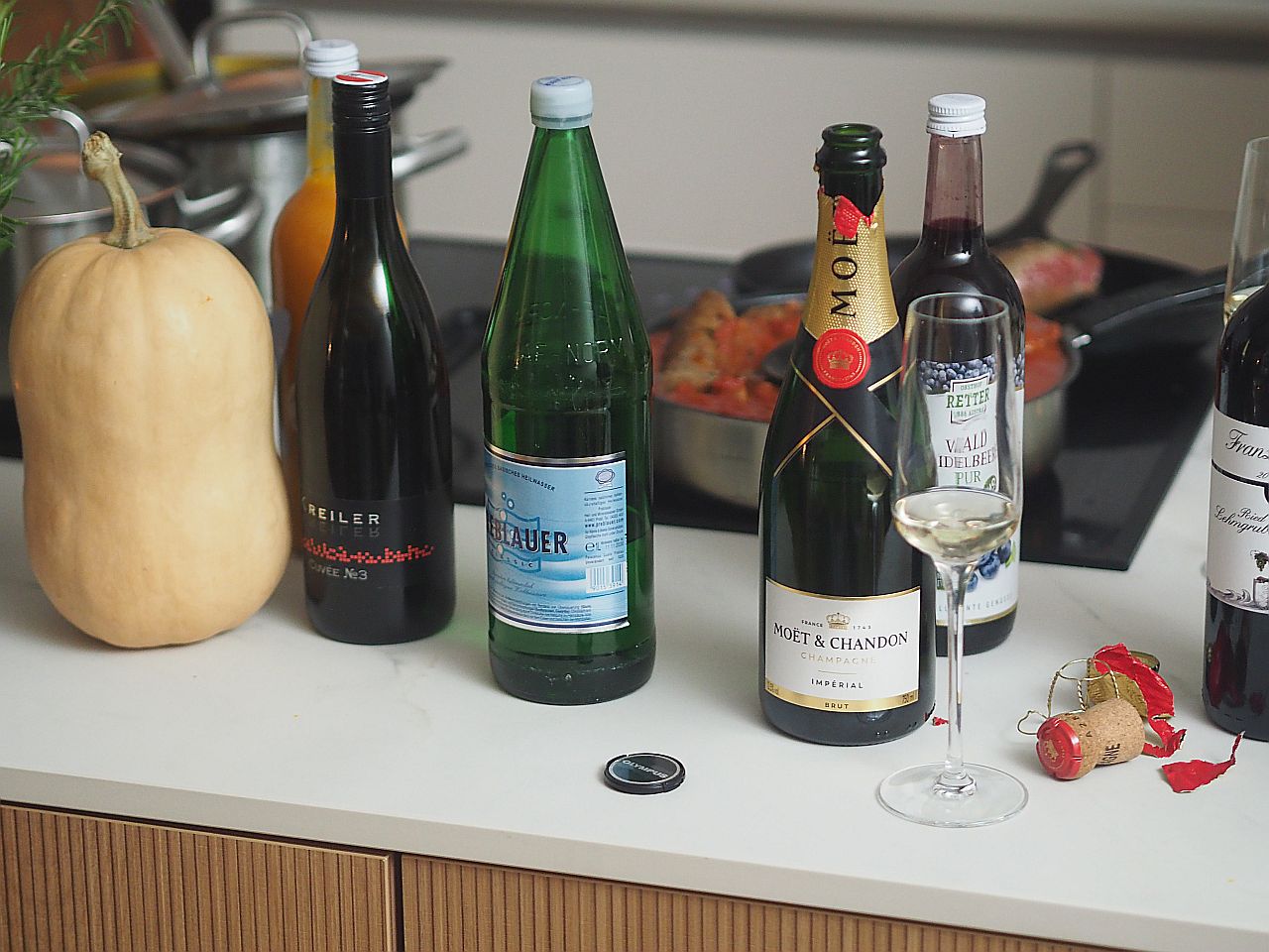 Eine Auswahl an Getränken auf einer Küchentheke, darunter ein Kürbis, Flaschen Mineralwasser, Wein und Champagner, sowie ein Glas mit Sekt und ein Korken.