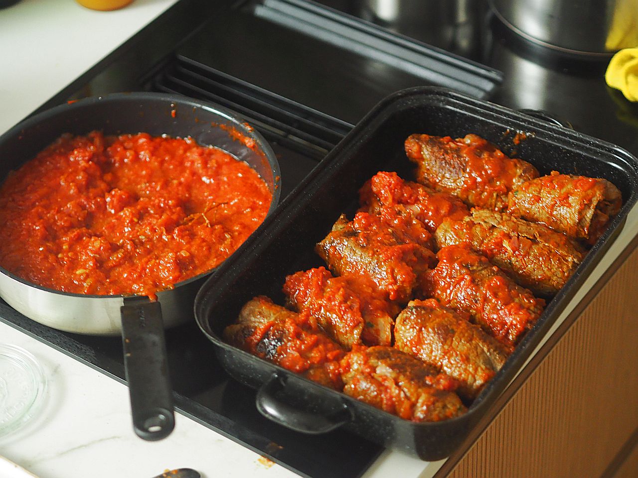 Ein Topf mit einer würzigen Tomatensoße und ein Blech mit gefüllten Rinderrouladen, die in der Soße serviert werden.