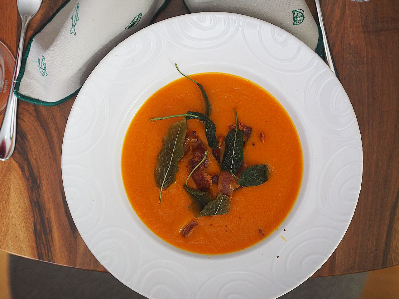 Eine cremige Kürbissuppe mit knusprigem Speck und frischen Salbeiblättern, serviert in einer weißen Schüssel auf einem Holztablett.