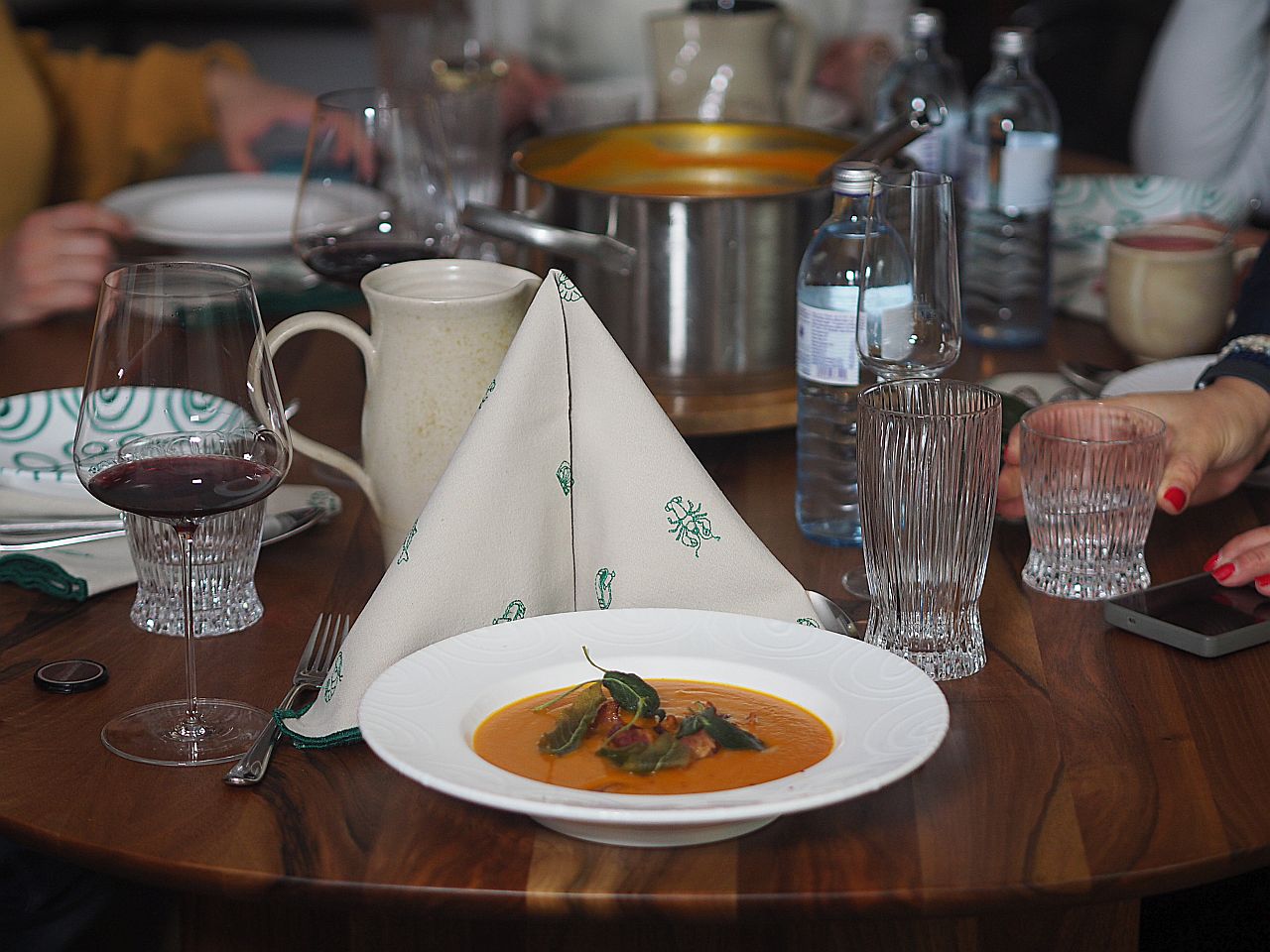Ein gedeckter Esstisch mit einer weißen Suppe in einer Schüssel, umgeben von Weingläsern, Wasserflaschen und Besteck. Im Hintergrund sind Hände zu sehen, die sich auf das Tischgedeck konzentrieren.
