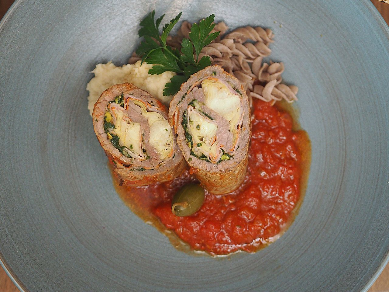 Ein Teller mit aufgeschnittenem Rinderroulade gefüllt mit Gemüse, serviert auf Tomatensauce und Beilage aus Püree und Vollkornnudeln.