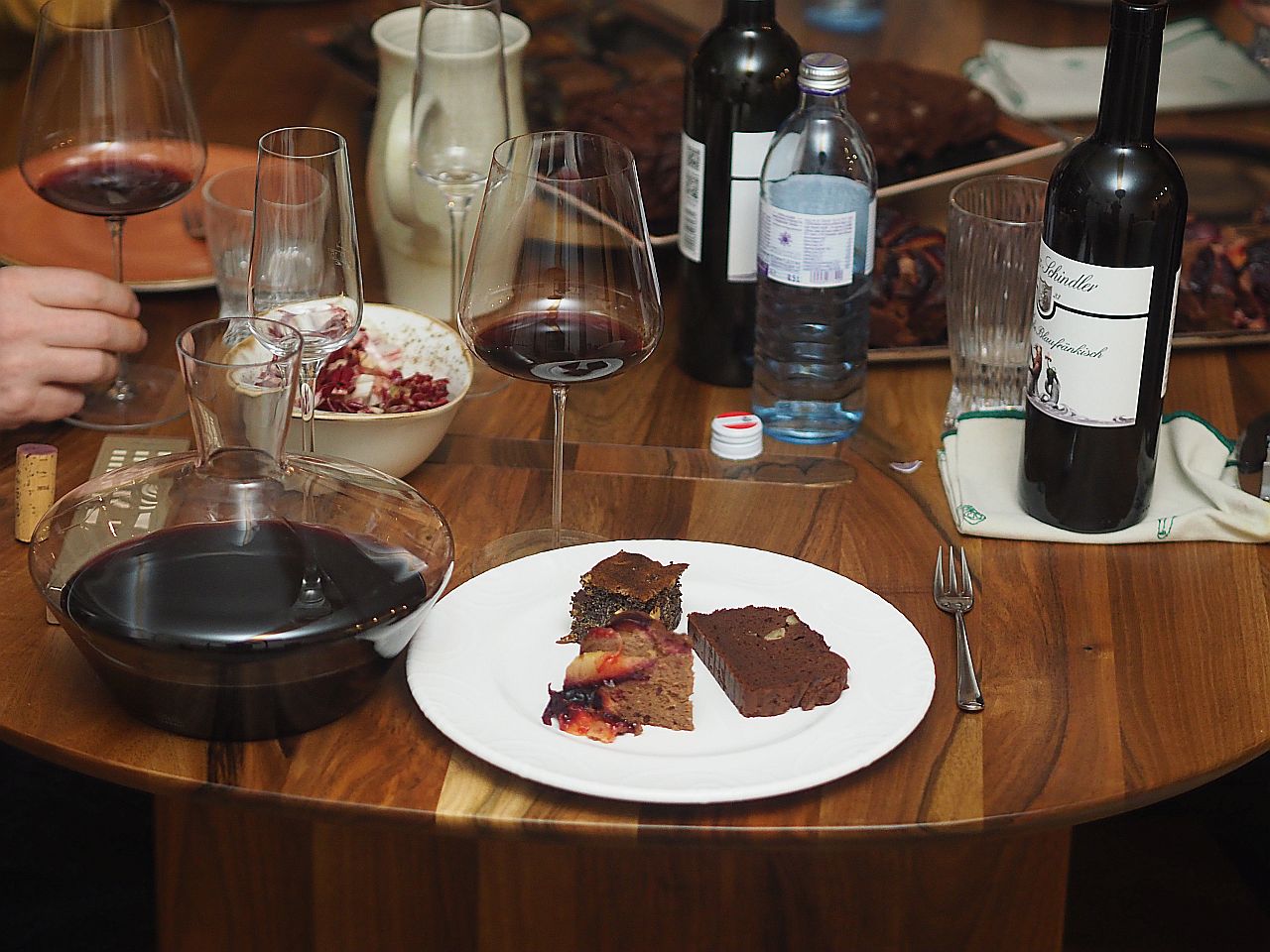 Ein Tisch mit verschiedenen Weinen, Wasserflaschen und einem Dessertteller mit zwei Stücken Kuchen. Eine Karaffe mit Rotwein steht ebenfalls auf dem Tisch.