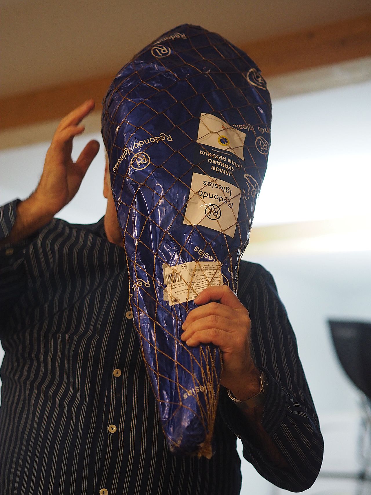 Eine Person hält einen großen, blauen, netzartigen Sack, der vermutlich einen luftgetrockneten Schinken enthält. Der Sack verdeckt teilweise das Gesicht der Person.