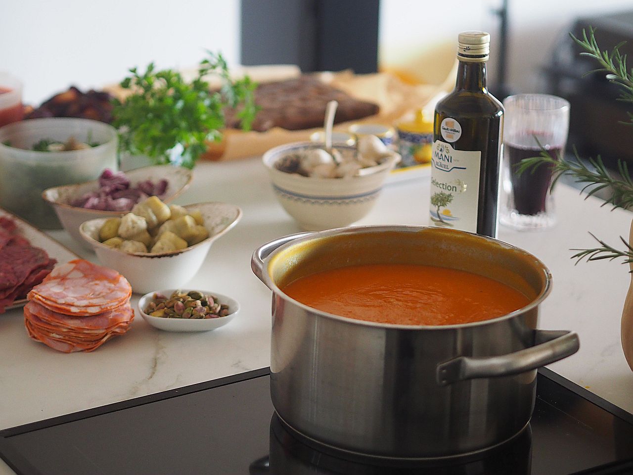 Ein Topf mit andersfarbiger Suppe steht auf einem Herd, umgeben von verschiedenen Zutaten wie Salami, Kräutern, eingelegtem Gemüse und Olivenöl auf einem Tisch.