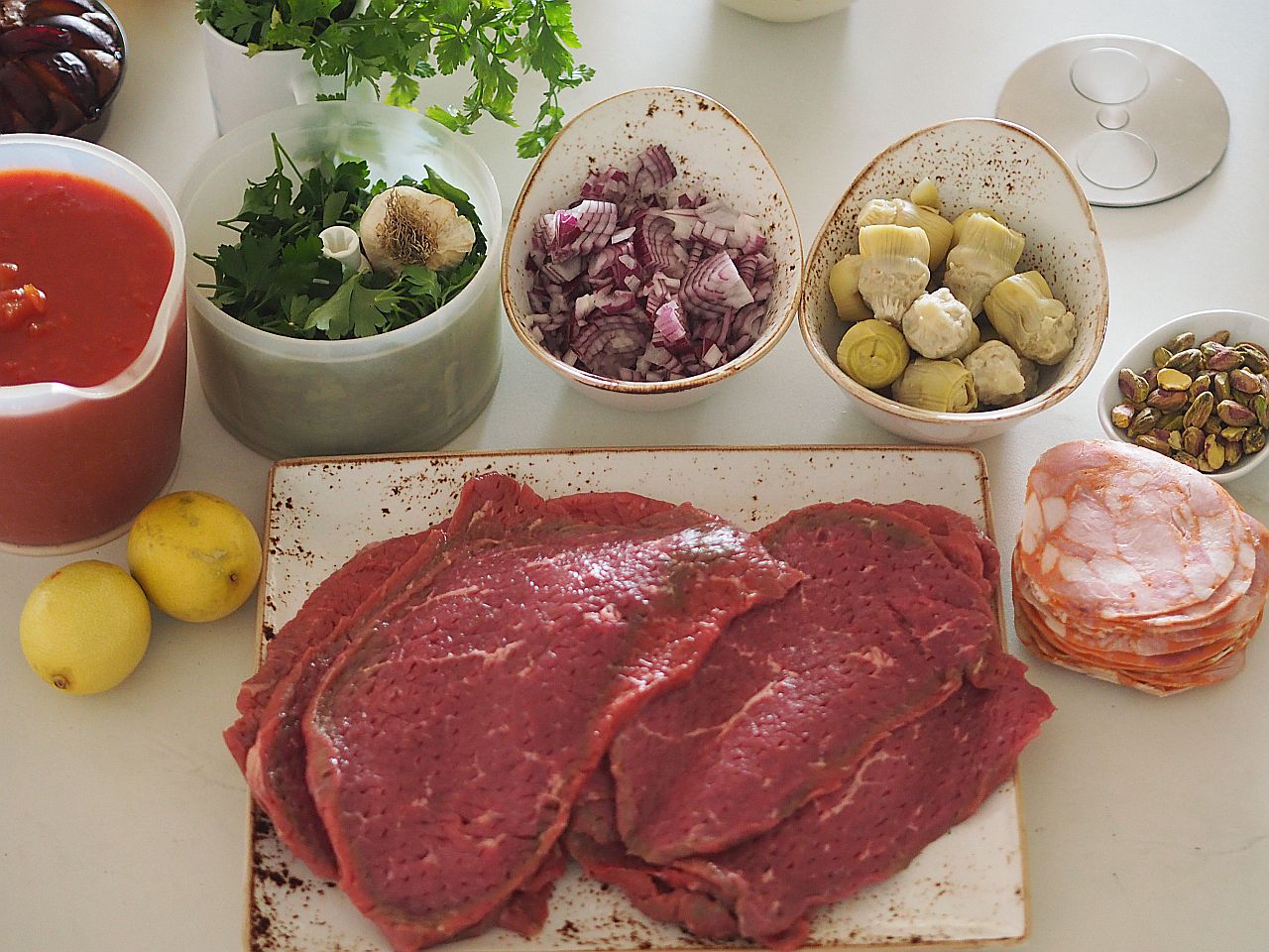 Eine Anordnung von Zutaten für ein Gericht: frisches rotes Fleisch, geschnittene Zwiebeln, Artischockenherzen, Koriander, Tomatensauce, Zitronen und Pistazien.