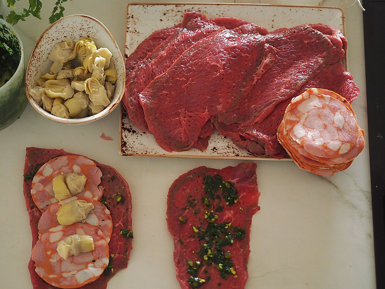 Rohes Fleisch auf einem Schneidebrett, daneben eine Schüssel mit Artischocken und einige Scheiben Wurst.