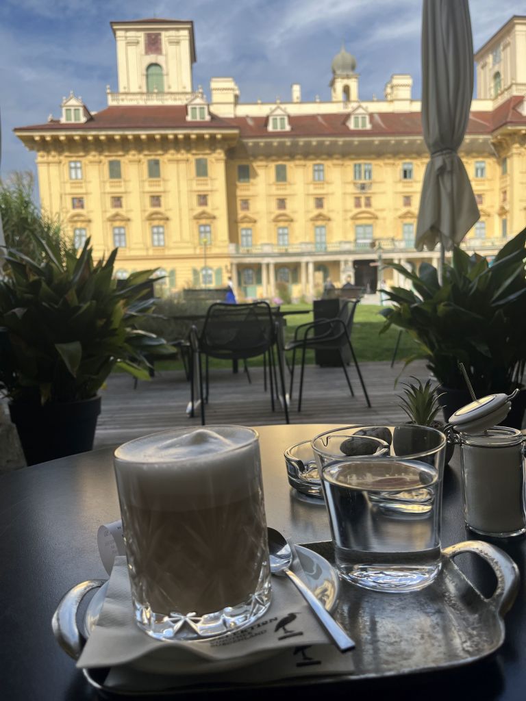 Ein Glas Latte macchiato und ein Glas Wasser auf einem Tisch, im Hintergrund ein gelbes Gebäude mit historischen Elementen und Pflanzen.