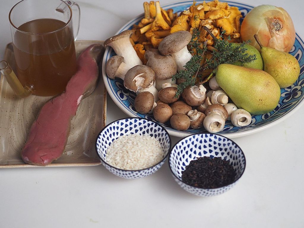 Ein Tablett mit einer Auswahl an frischen Zutaten: eine Tasse Brühe, eine Scheibe Fleisch, verschiedene Pilze, zwei Birnen, eine Zwiebel, etwas frischer Thymian, sowie zwei kleine Schalen mit Reis und schwarzem Reis.