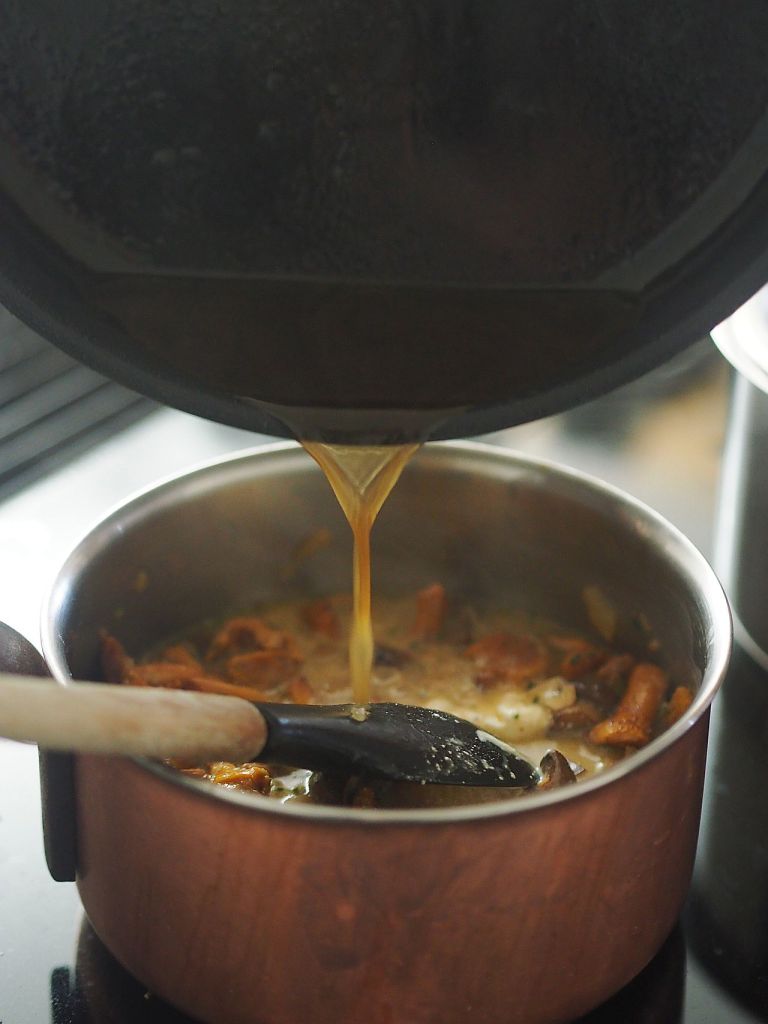 Ein Topf mit einer goldenen Brühe, die aus einem anderen Gefäß gegossen wird, während ein Holzlöffel im Topf liegt.