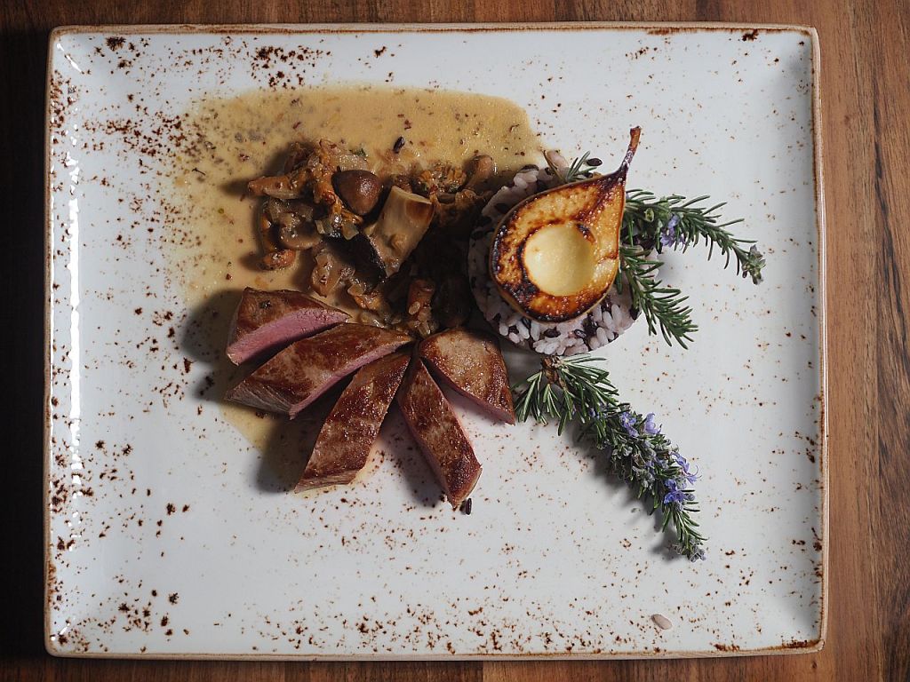Ein kunstvoll angerichteter Teller mit zartem Fleisch, einer cremigen Sauce, Pilzen, Reis und einer gebratenen Birne, garniert mit frischen Kräutern.