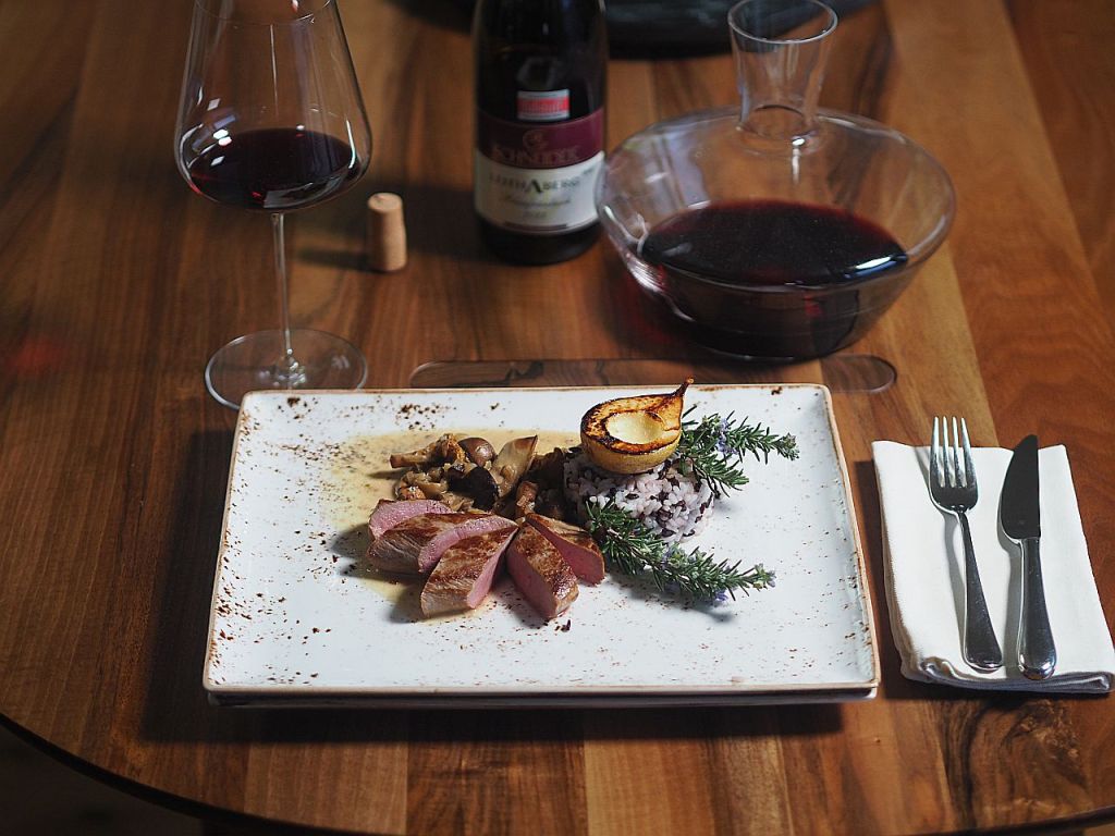 Ein elegant angerichteter Teller mit gebratenem Fleisch, Beilagen aus Pilzen und schwarzem Reis, garniert mit einem gegrillten Stück Zitrone und frischen Kräutern. Im Hintergrund ein Glas Rotwein und eine Dekantierkaraffe.