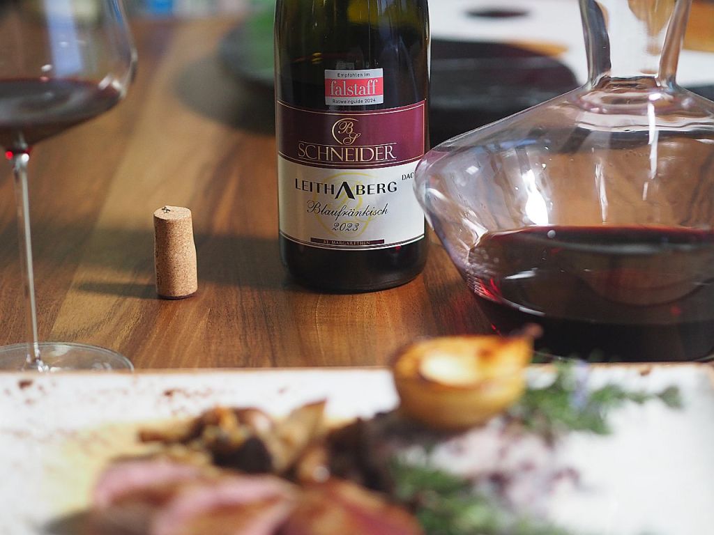 Ein Glas Rotwein, eine Flasche Schneider Leithaberg Blaufränkisch 2023 und eine Karaffe auf einem Holztisch, daneben ein Korken und ein Teller mit ansprechendem Essen.