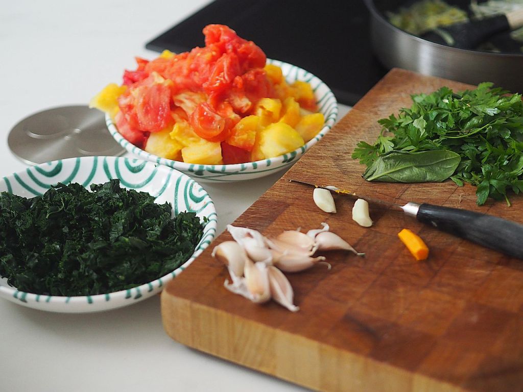 Vorbereitete Zutaten für ein Rezept, einschließlich gehacktem Grünkohl, gewürfelten Tomaten und Ananas, frischem Koriander, Knoblauchzehen und einem Stück Ingwer auf einem Holzbrett.