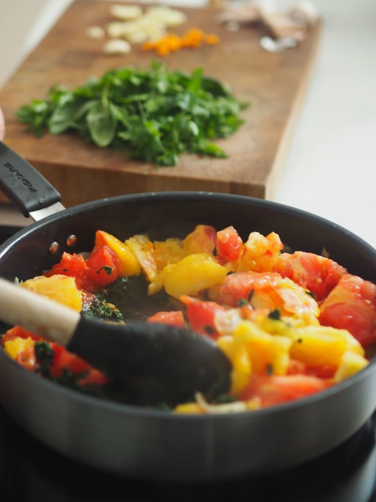Eine Pfanne mit buntem Gemüse, darunter Tomaten und Ananas, die auf dem Herd gekocht werden. Im Hintergrund frische Kräuter und geschnittene Zutaten auf einem Schneidebrett.