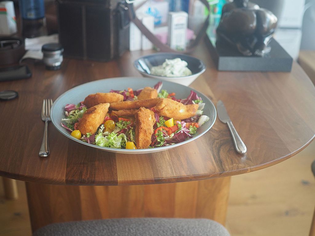 Ein Teller mit paniertem Hühnchen auf einem bunten Salat, serviert auf einem Holztisch, mit Besteck neben dem Teller.