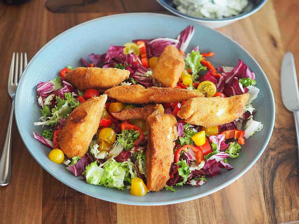 Ein Teller mit knusprig gebratenen Fischfilets auf einem bunten Salat aus frischem Gemüse wie Rot- und Grüner Salat, Paprika und Kirschtomaten.